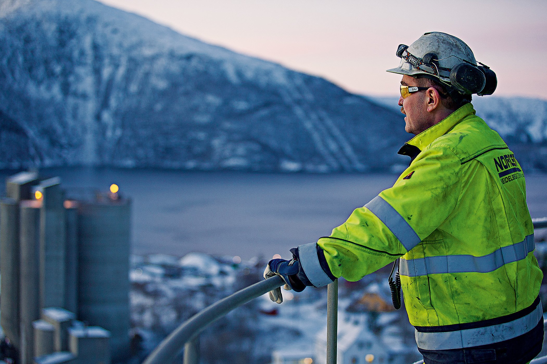 A man with a yellow vest is looking at the cement plant below