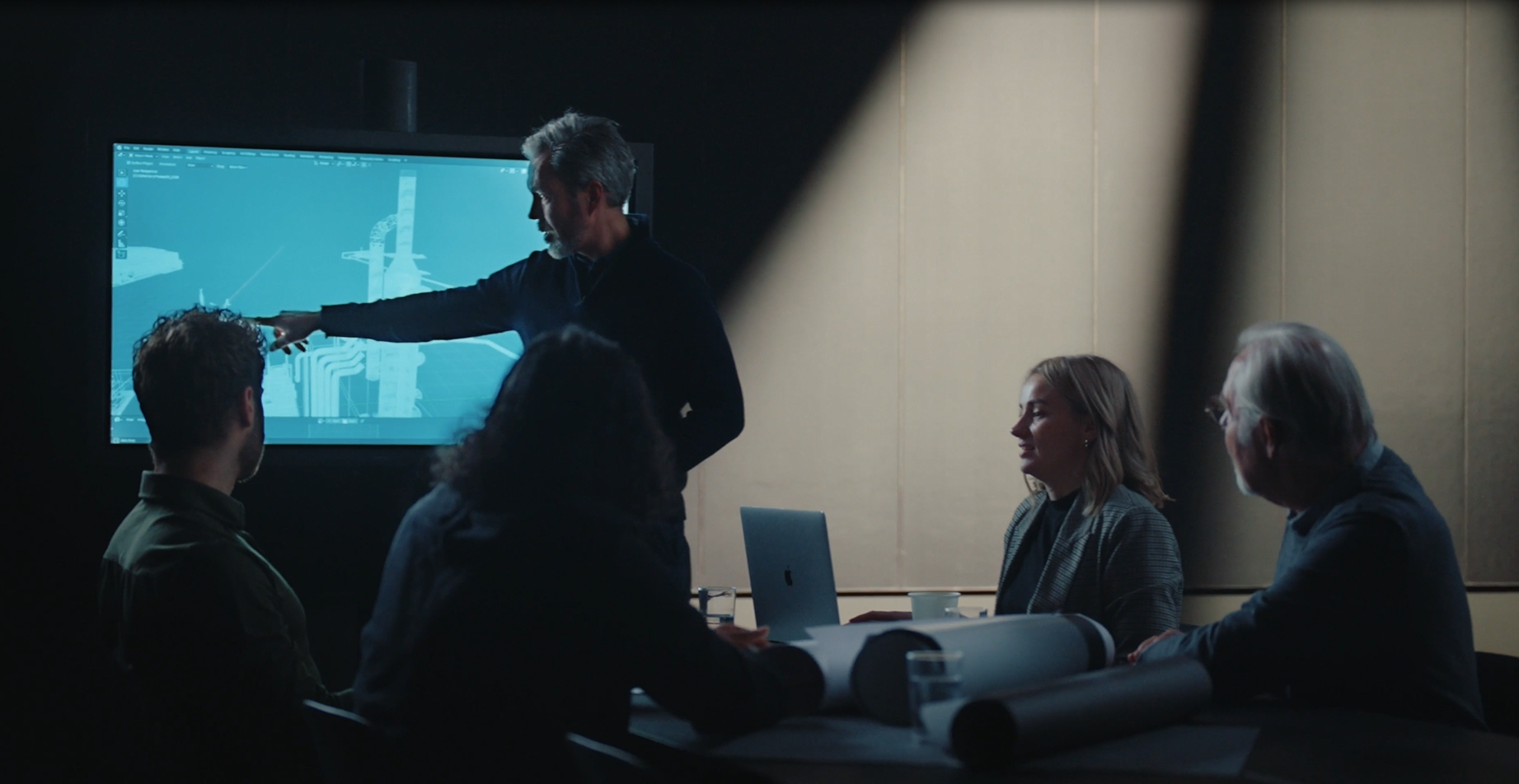 A man points to a monitor in a darkened room to explain something to the other people present