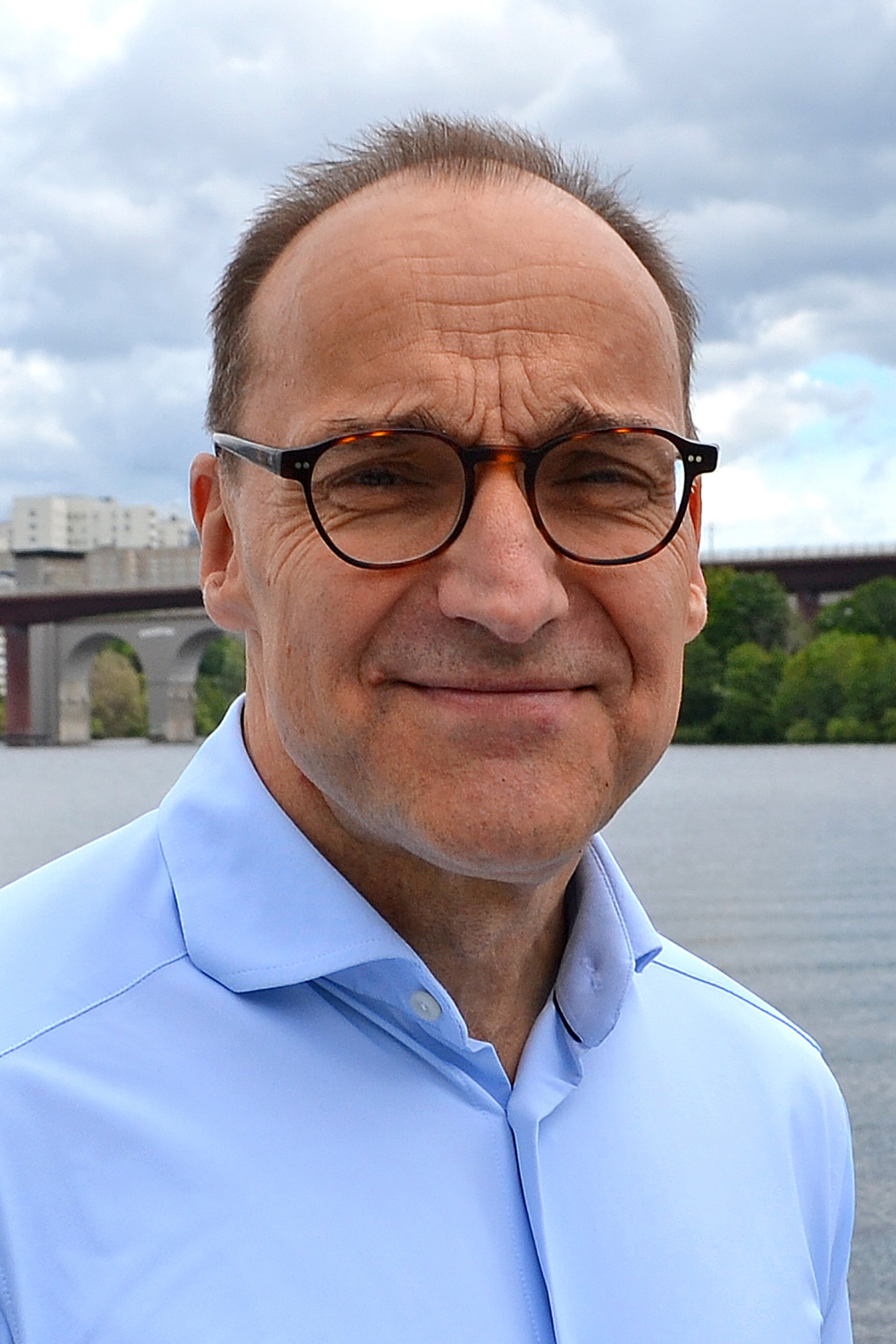 A person wearing a light blue shirt is standing near a body of water, with a cloudy sky and a bridge in the background surrounded by greenery.