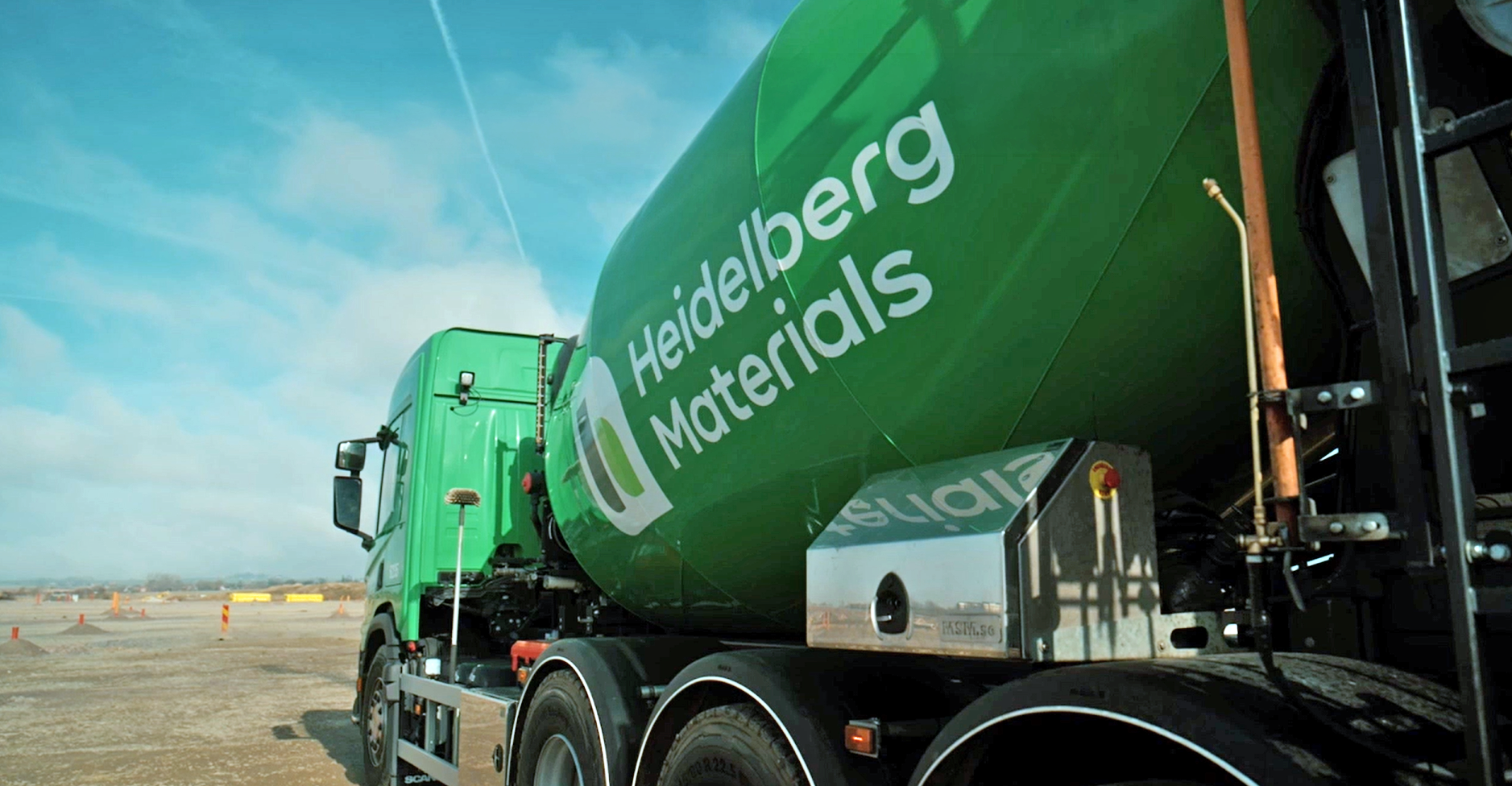 A green Heidelberg Materials concrete mixer truck parked on an industrial site under a partly cloudy sky.