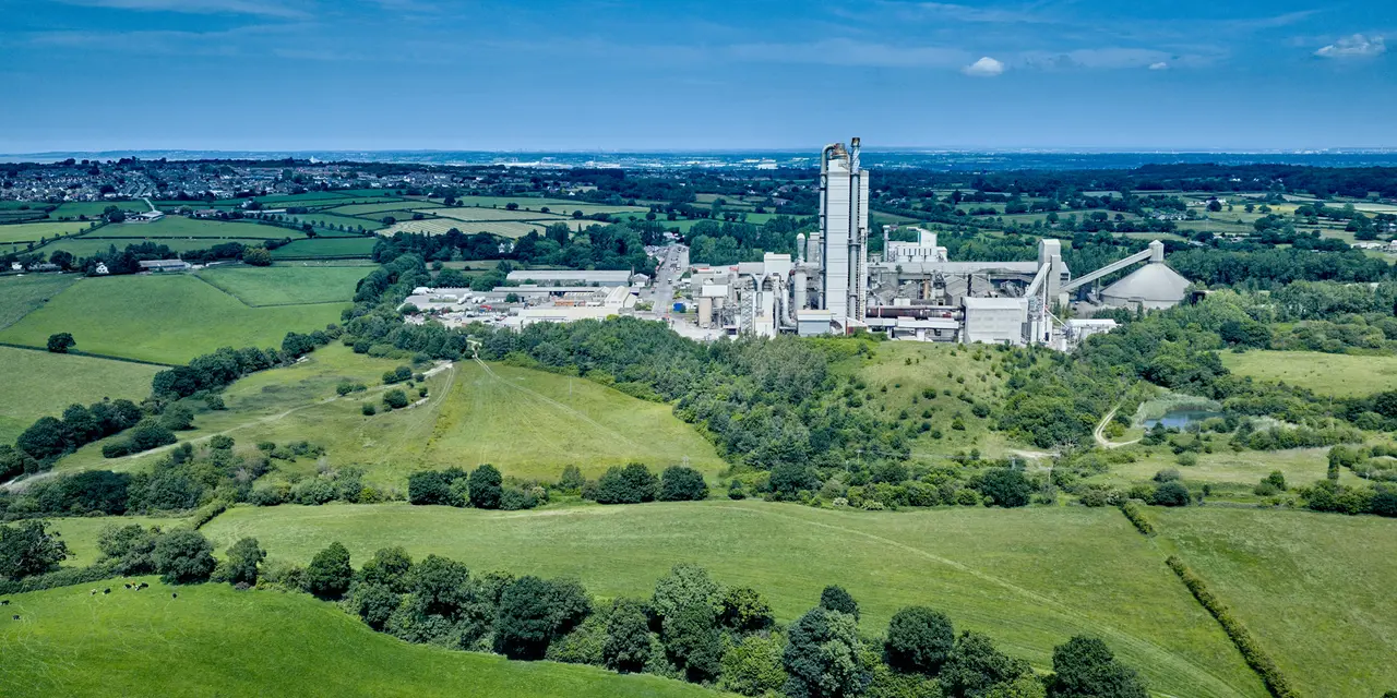 Aerial view of a large cement factory surrounded by green fields and trees, with industrial buildings and tall chimneys at the center. Rolling countryside extends into the background under a clear blue sky.
